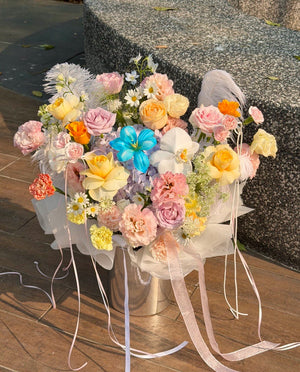 A large, vibrant floral bouquet in a silver metal box, decorated with pastel pink, yellow, peach, and blue flowers, white daisies, and white feathers, tied with long sheer pink and white ribbons, set outdoors on a wooden deck next to a stone bench.