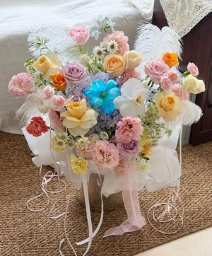 A pastel floral arrangement with buttercup roses, champagne roses, pink roses, filler and purple hydrangea placed in a silver box.