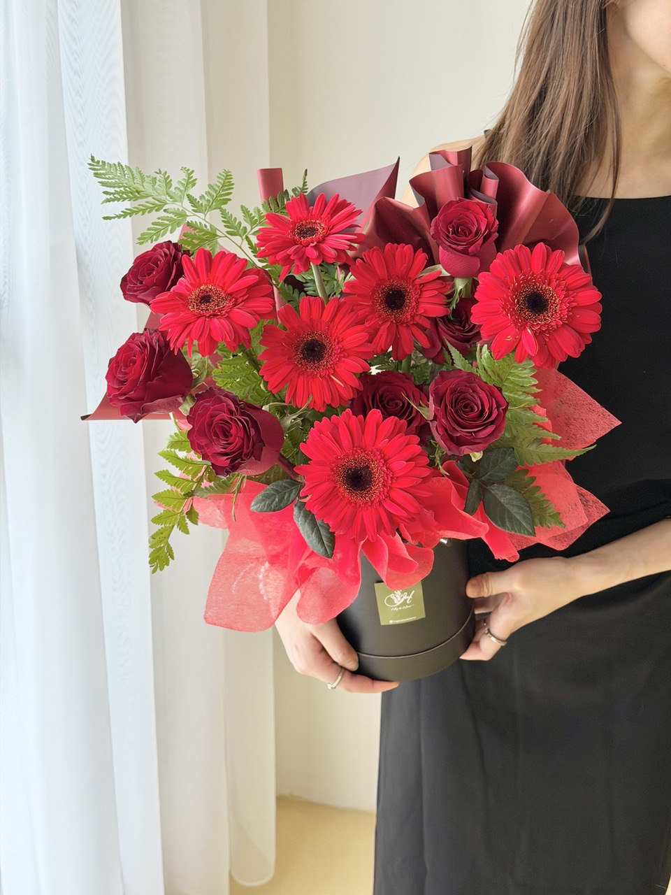 Bouquet of red gerbera and red roses with greenery in a black box held by a person against a white curtain at Subang Jaya, perfect for house warming or grand opening flower stand.