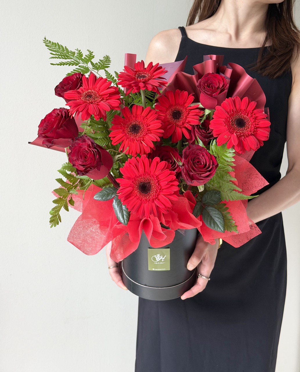 Person holding a bouquet of red gerbera and red roses in a black box against a neutral background.