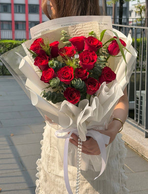 A person outdoors holding a large bouquet of red roses wrapped in white paper with a white ribbon and pearl-like hanging detail. The background shows a paved walkway, metal railing, and buildings.