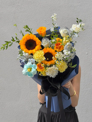 Bouquet of sunflowers with dye tulips, refulgence rose, white rose, white matthiola, yellow carnation and chamomile held by a person against a gray background for graduation.