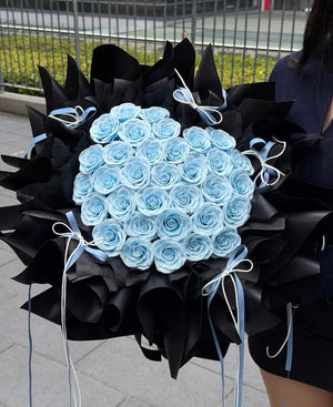 A large bouquet of light blue soap flower roses wrapped in black paper with pale blue ribbons, held by a person outdoors near a metal fence and paved ground.