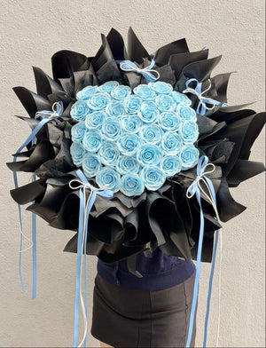 A large bouquet of light blue soap flower roses wrapped in black paper with light blue ribbons, held by a person wearing a navy top and dark skirt against a plain light gray wall.
