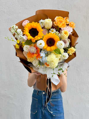 Bouquet of sunflowers and pink rose, dancing lady and other fillers wrapped in brown paper held by a person against a plain background.