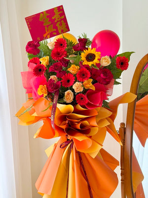 A vibrant business opening flower stand with red, pink, yellow, and orange flowers, a red balloon, and a red Congratulations card, wrapped in orange paper and tied with a ribbon, set next to a wooden mirror.