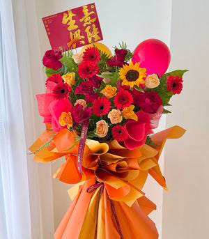 A vibrant business opening flower stand with red, pink, yellow, and orange roses, a large pink balloon, and orange paper wrapping, featuring a red congratulation card.