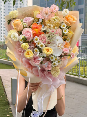 A person outdoors holding a large, vibrant bouquet of fresh flowers. The bouquet features a mix of pastel pink, yellow, orange, and white flowers wrapped in beige paper with white ribbons.