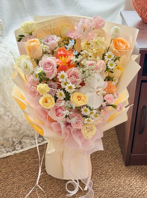 A large, vibrant floral bouquet wrapped in pale pink and cream paper, filled with roses, daisies, and other small flowers, set on a carpeted floor next to a wooden cabinet.