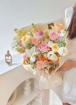 A spring-themed bouquet of assorted flowers in shades of pastel pink, yellow, white, and blue, held beside a woman in a white lace dress.