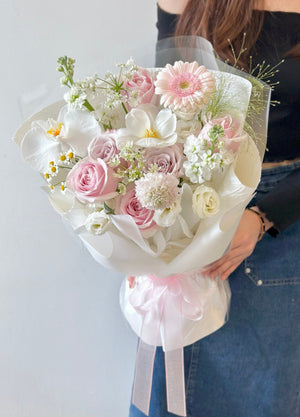 A woman in a black off-shoulder top and denim skirt holding a pastel-colored bouquet of roses, gerbera, and phalaenopsis wrapped in white paper with a pink ribbon.
