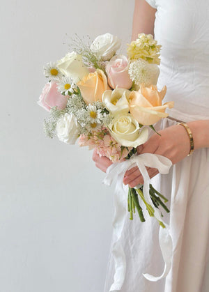 Elegant pastel wedding bouquet featuring champagne rose, white rose, hana white, matthiola, white tulip tied with a flowing white ribbon—perfect for bridal or romantic occasions