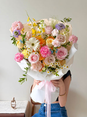 Pastel mixed flower bouquet with pink roses, carnations, gerbera daisies and chamomile wrapped in white paper against a plain background.