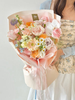A woman in a floral corset top and white skirt holds a pastel-colored bouquet featuring roses, hana white, carnations, quicksand rose, gerbera, menta rose, phalaenopsis and orange tulip wrapped in pink and cream paper.