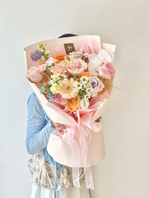 Delicate floral arrangement with roses, hana white, carnations, quicksand rose, gerbera, menta rose, phalaenopsis and orange tulip wrapped in pink and cream paper, wrapped in peach and ivory paper, held by a woman in floral attire.