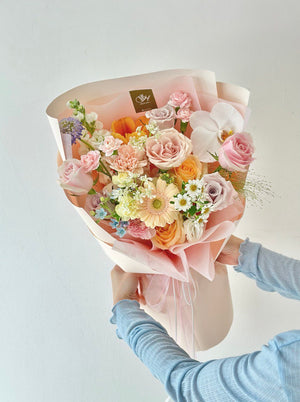 Elegant bouquet of mixed fresh flowers in soft pink, yellow, peach, and lavender tones, wrapped in ribbon-tied paper, being held by a woman.