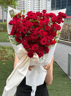 A large heart-shaped bouquet of deep red roses rose spray wrapped in white paper with a white ribbon bow, held by a person standing outdoors on a paved walkway with trees and modern buildings in the background.