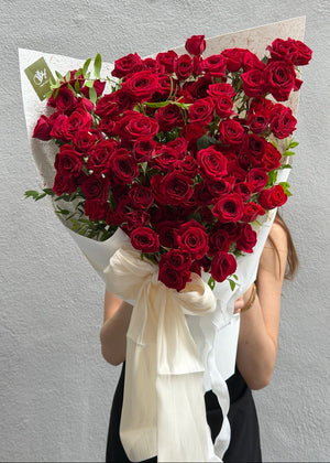 A large bouquet of deep red rose spray wrapped in white paper with a cream ribbon bow, held in front of a person against a light gray textured wall.