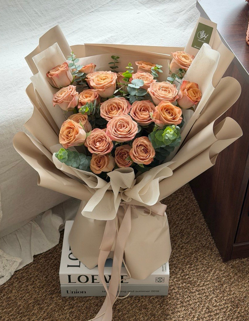 A bouquet of cappuccino roses wrapped in beige paper with a ribbon bow, placed on top of a book on a carpeted floor next to a sofa.