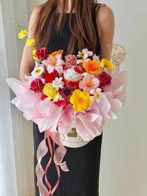 A person in a black sleeveless dress holds a colorful soap flower with red lucky cat wrapped in light pink wrapper paper. The bouquet includes yellow, orange, red, and pink flowers, a white cat figurine, and pink ribbon ties, set against a plain indoor background.