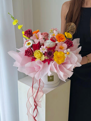 A vibrant, multicolored floral arrangement in a white box, wrapped in light pink tissue paper and tied with a pink ribbon. The bouquet features a white maneki-neko (lucky cat) figurine, yellow orchids, red and yellow roses, and white daisies.