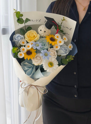A person in a dark navy shirt holds a beautifully arranged bouquet of soap flowers, including yellow roses, light blue roses, daisies, and a small white plush bear wearing a graduation cap, wrapped in cream paper with a brown twine bow.