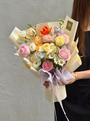 A person holding a large, beautifully arranged floral bouquet with pink, yellow, peach, and white flowers wrapped in cream paper with lavender ribbons, set against a neutral gray wall.
