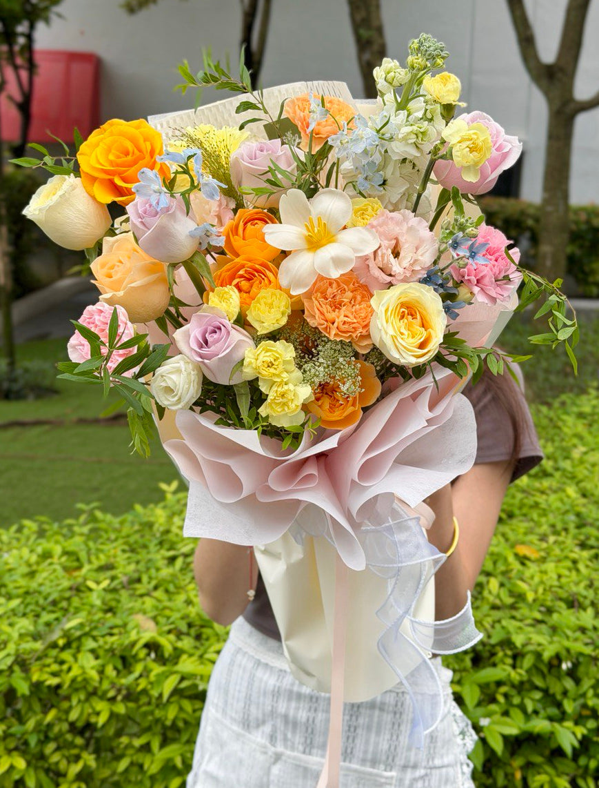 Colourful mixed flower bouquet with buttercuproses, refulgence roses, pastel carnations and seasonal fillers, hand-wrapped in soft cream paper.