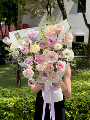 A purple theme flower bouquet with purple tulips, purple carnation, menta roses, lilac roses and some lace flowers is held by a girl outdoors with greenery in the background.