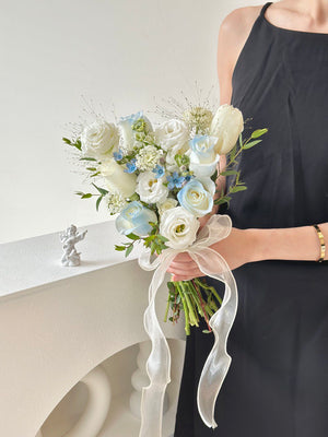 Elegant ROM bouquet featuring white and soft blue blooms, tied with a flowing sheer ribbon and held in a modern minimalist setting.