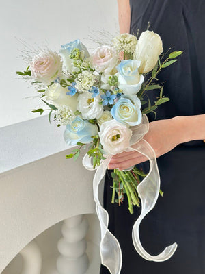Elegant ROM bouquet with white and baby blue flowers, tied with a sheer ribbon, held by a woman in a black dress beside a minimalist white table.