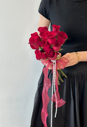 A romantic ROM bouquet of deep red roses tied with a sheer burgundy ribbon and cascading pearl strands, held by a woman in a black dress.