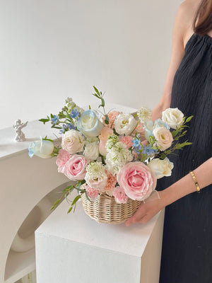 A woman presents a basket of flowers, including pink and blue roses, in front of a white table conveying a get well soon message.