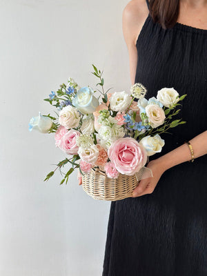 A woman stands infront of a wall, holding a flower basket filled with pink and blue roses and assorted soft-colored blooms.