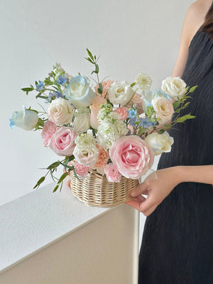 A woman holds a basket of pink and blue roses, along with mixed soft-colored flowers, in front of a window.