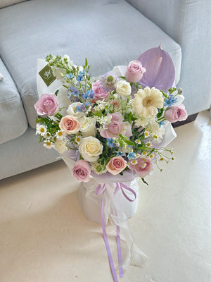 Charming birthday bouquet with a blend of blush roses, white gerberas, and mixed flowers, wrapped in white and tied with lavender ribbons placing on floor.