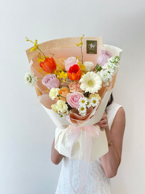 Delicate spring bouquet placed on a white pedestal, featuring a harmonious blend of tulips, roses, carnations, and filler flowers against a minimalist backdrop.