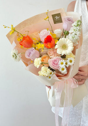 A vibrant bouquet named "Spring Serenade" featuring a mix of orange tulips, champagne roses, white gerbera, and hana white wrapped in peach-toned paper with a pink ribbon.