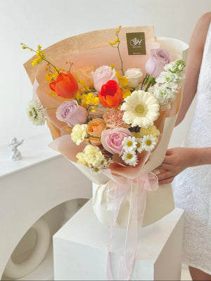 Elegant floral arrangement with orange tulips, soft pink and lilac roses, creamy carnations, yellow dancing orchids, and white daisies held by a woman in a white eyelet dress.