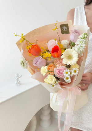A beautifully wrapped bouquet with fresh seasonal flowers in shades of orange, blush, yellow, and white, tied with sheer pink ribbon, exuding a soft romantic charm.