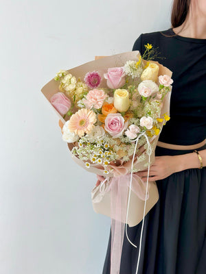 A pastel-toned flower bouquet featuring pink roses, peach gerbera, chamomile, and cream-colored blooms wrapped in soft beige paper.