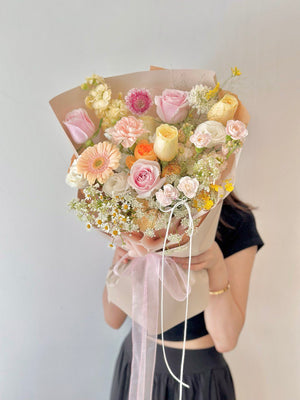 Elegant hand bouquet with soft pink, cream, and peach flowers, tied with a light pink ribbon and held by a woman in a black dress.