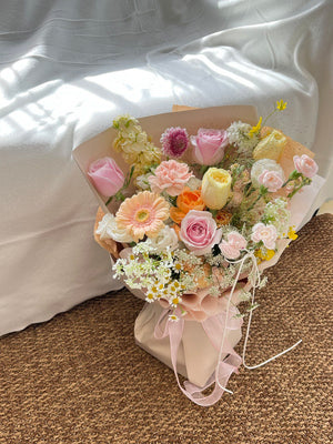 A pastel-toned flower bouquet featuring pink roses, peach gerbera, chamomile, and cream-colored blooms wrapped in soft beige paper.