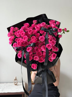 Bouquet of hot pink rose spray wrapped in black paper with a black ribbon on a white background.