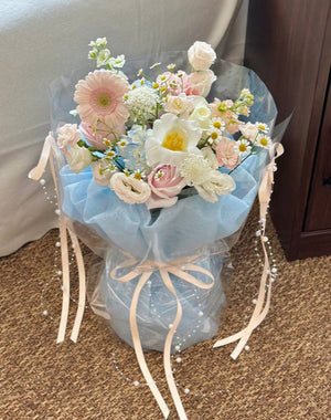 A soft pastel flower bouquet wrapped in sheer plastic and baby blue fabric, featuring blush pink roses, white tulip, pink gerbera daisies, chamomile, and delicate filler flowers, adorned with cream-colored ribbons and pearl strands.