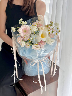 A pastel-toned flower bouquet held by a woman in a black dress, featuring soft pink gerberas, white roses, white tulip, chamomile, and carnation, wrapped in clear plastic with baby blue fabric and cream pearl ribbon.