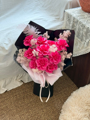 A bouquet of pink soap flowers arranged in a black box with pale pink tissue paper and a white feather, photographed on a carpeted floor beside a bed and a lace-covered table.