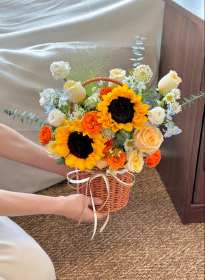 Person holding a basket of flowers with sunflowers, buttercup rose, hana white, delphinium blue and eustoma on a brown carpeted floor.