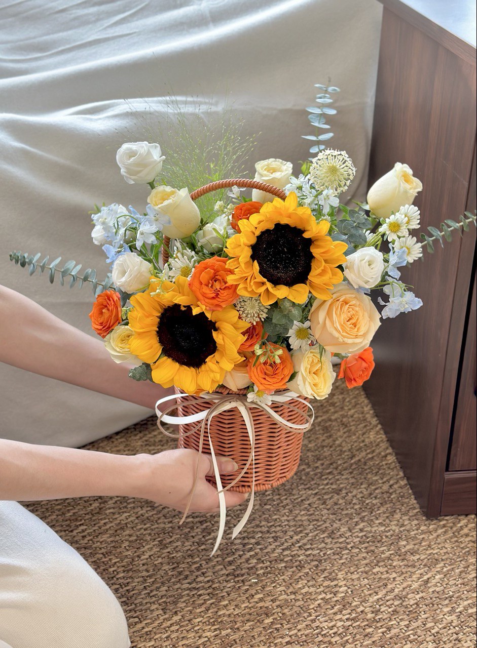 Person holding a basket of flowers with sunflowers, buttercup rose, hana white, delphinium blue and eustoma on a brown carpeted floor.
