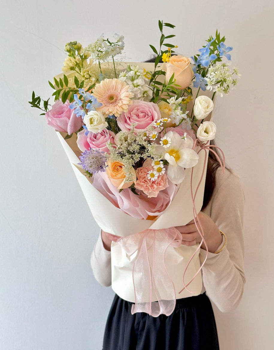 Person holding a bouquet of flowers arrangement of pastel roses, tulip, and delphinium blue in a modern light yellow wrap stand against a plain background
